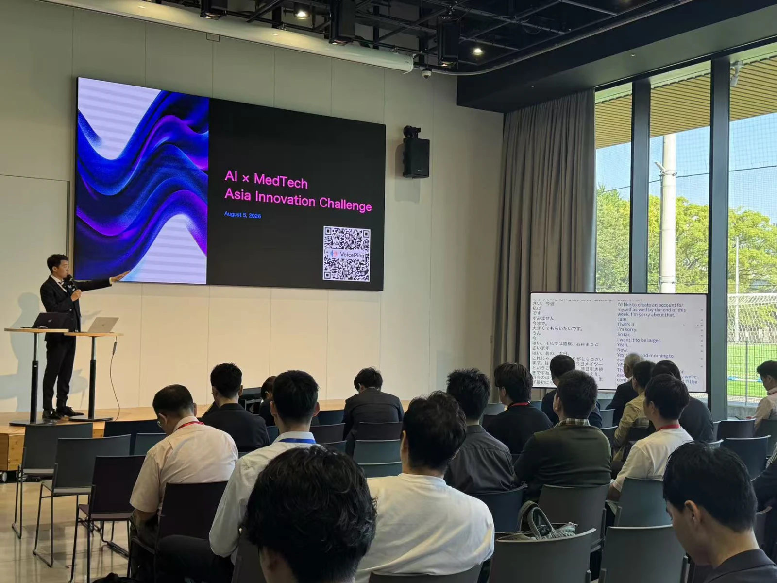A speaker introduces AI×MedTech at STATION Ai, with the event title, a VoicePing QR code, and Japanese–English captions visible beside the stage.
