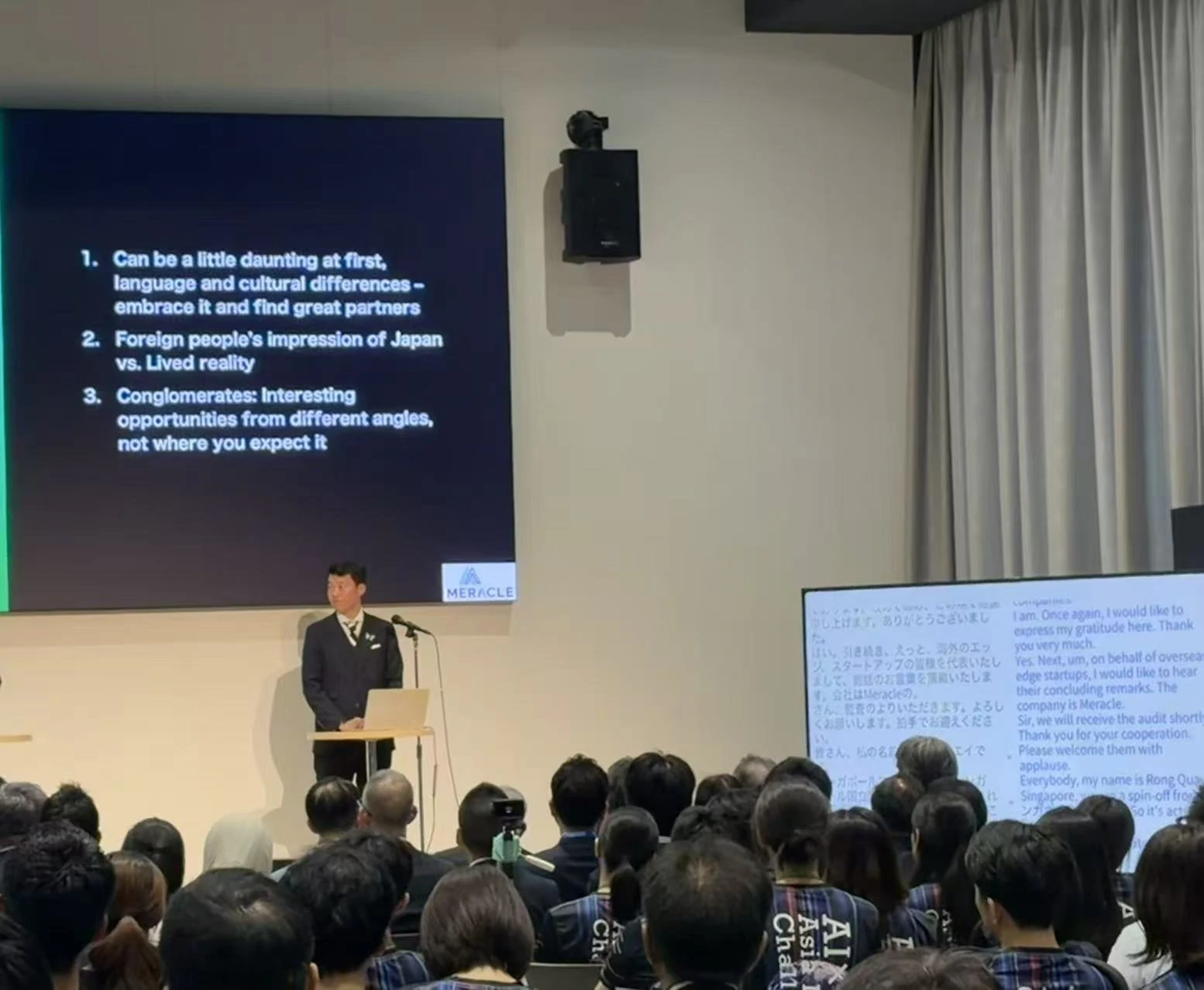 An AI×MedTech speaker presenting beside a Japanese–English live caption display at STATION Ai.