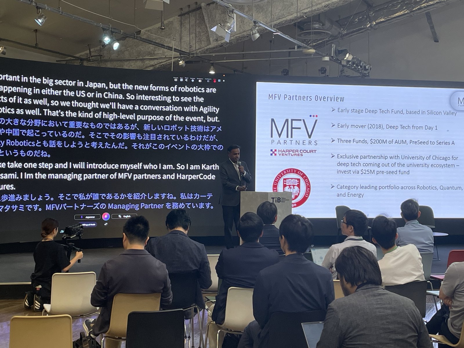 Near the podium, the speaker presents an overview slide while VoicePing’s live English and Japanese translation scrolls on the screen beside it.