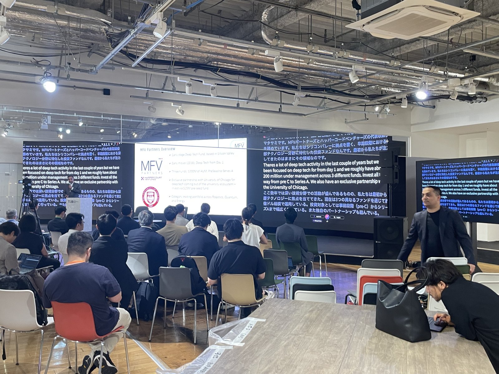 A wide view of the room at Tokyo Innovation Base: attendees seated toward two large screens, one carrying the presenter’s slides and the other running VoicePing’s live English-Japanese translation.