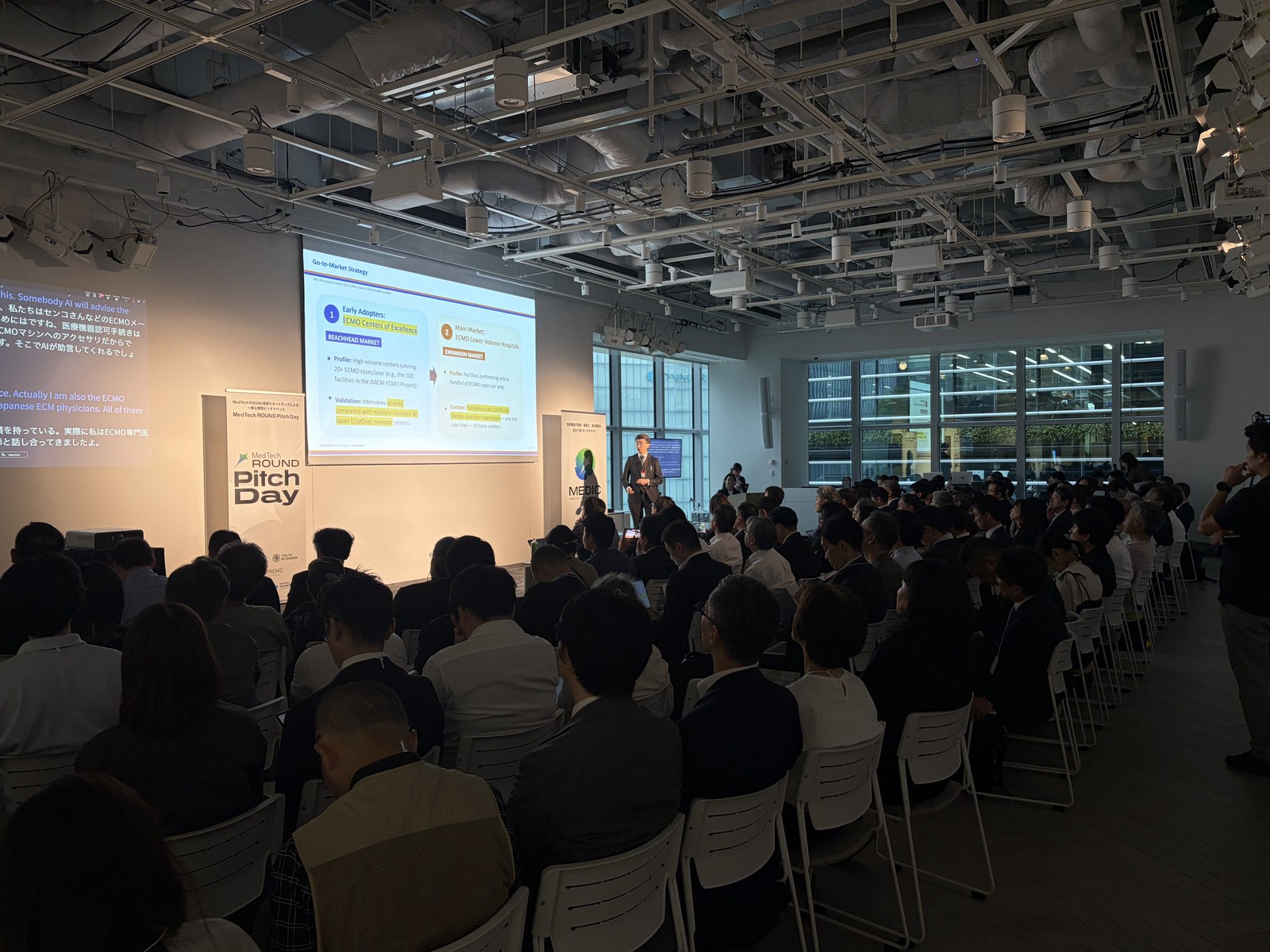 A startup pitches in English on the main stage at MedTech ROUND Pitch Day, while a second screen beside it shows the talk translated live into Japanese.